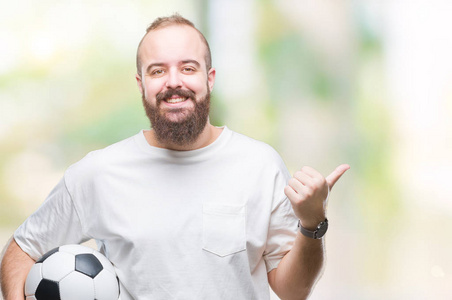 年轻的白种人潮人拿着足球球在孤立的背景上指指点点，并用拇指向旁边露出快乐的脸微笑