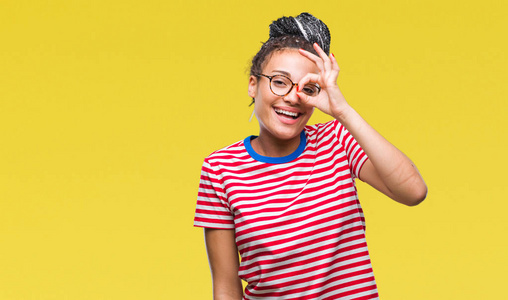 年轻的辫子非洲裔美国女孩戴着眼镜在孤立的背景上做着好的手势，用手微笑的眼睛透过手指看着快乐的脸。