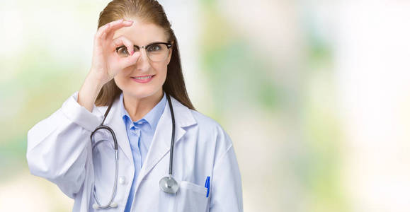 中年成熟的医生女人穿着医用外套，在孤立的背景下做着OK手势，用手微笑，用快乐的脸透过手指看。