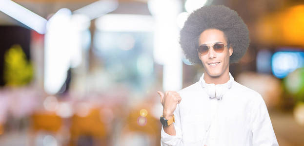 年轻的非洲裔美国男子带着非洲头发，戴着太阳镜和耳机，微笑着，快乐的脸看着，拇指向上指向一边。