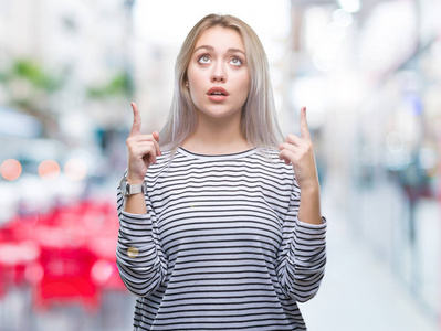 年轻的金发女人在孤立的背景上惊讶和惊讶地抬头，手指和举起的手臂。