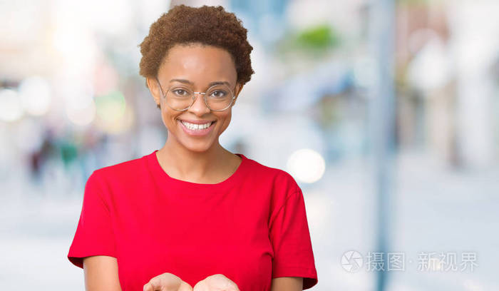 美丽的年轻非洲裔美国妇女戴着眼镜，在孤立的背景上微笑，双手掌心一起接受或给予手势。 持有和保护