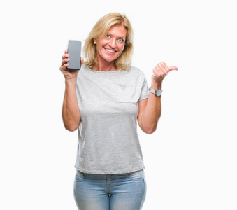 中年金发女性，在孤立的背景上显示智能手机的空白屏幕，用拇指指向侧面，脸上洋溢着幸福的笑容