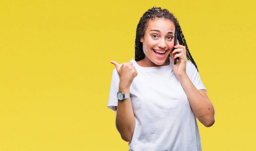年轻的辫子非洲裔美国女孩显示使用智能手机在孤立的背景上打电话，用拇指指向一边，脸上洋溢着幸福的笑容