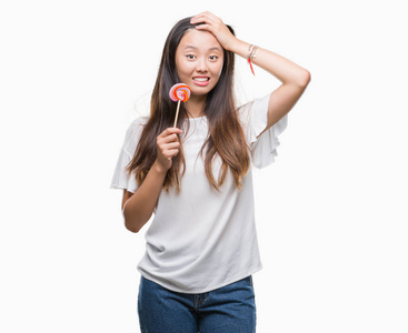 年轻的亚洲女人吃棒棒糖在孤立的背景下强调，手在头上震惊，羞愧和惊讶的脸，愤怒和沮丧。 因为错误而恐惧和不安。