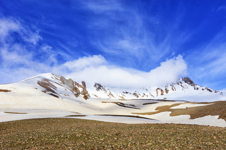 在阳光明媚的春天，土耳其中部的埃尔西伊斯山的雪顶被白云笼罩在明亮的蓝天上。