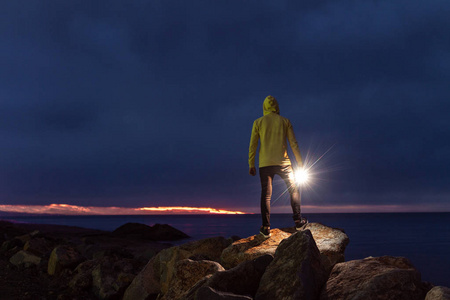 戴着引擎盖的人站在岩石上，手里拿着手电筒，在晚上的海洋背景上。 平静宁静孤独与自然。