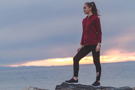 健身迷人的女人穿着运动鞋，在锻炼后爬上岩石，并在日落时在海上背景上眺望远方。 运动和积极的生活方式