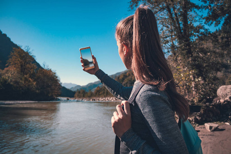 年轻着蓝色背包的年轻女孩游客在山上拍摄河流的照片。 旅游享受美丽的风景和自然