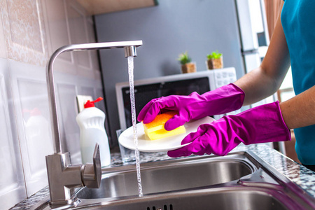 家庭主妇戴着橡胶彩色手套，在家里的厨房里用黄色海绵洗碗。