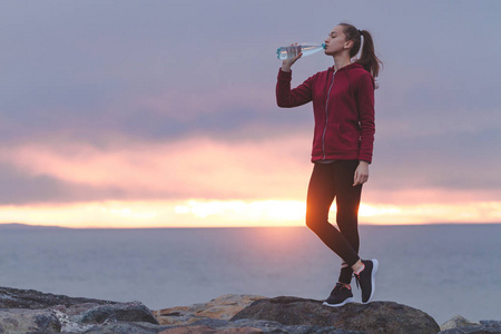 健身有吸引力的女人穿着运动鞋站在石头上喝水后,在日落的海上背景下