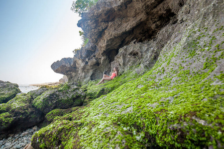 印度尼西亚巴厘岛的绿碗海滩上，一位长发的年轻女子穿着粉红色的泳衣躺在岩石上，海洋附近有绿色的植物。