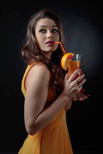 年轻漂亮的女孩穿着一件橙色的裙子，妆容完美，拿着一杯鸡尾酒。 鸡尾酒龙舌兰日出装饰着樱桃和一片橘子。