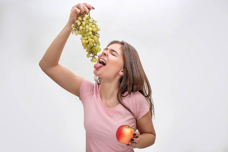 年轻女子拿着葡萄的照片打开了嘴。她用舌头伸手去。苹果在另一只手。隔离在灰色背景上