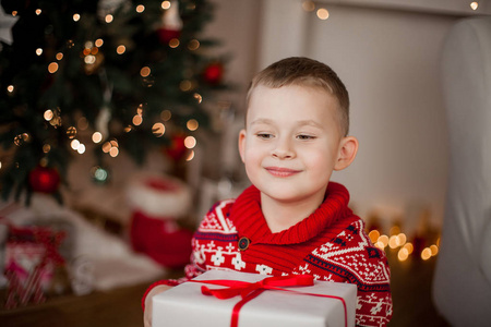 穿着红色圣诞毛衣的小可爱男孩坐在圣诞树附近。 一个4岁的快乐男孩的肖像和一个礼品盒。 假日。 圣诞节。 新年。