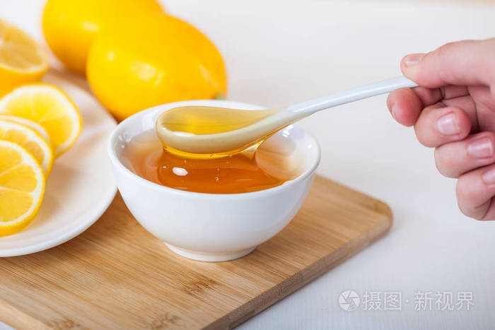 蜂蜜放在白色陶瓷碗里,勺子和柠檬放在木制厨房板上.