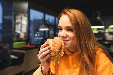 特写一个女孩的肖像，手里拿着一个美味的汉堡，笑着。 一个积极的青少年女孩晚上在一家舒适的餐馆吃快餐。
