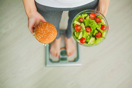 饮食。妇女测量体重在称重秤持有汉堡和沙拉。糖果是不健康的垃圾食品。节食, 健康饮食, 生活方式。减肥。肥胖。顶视图