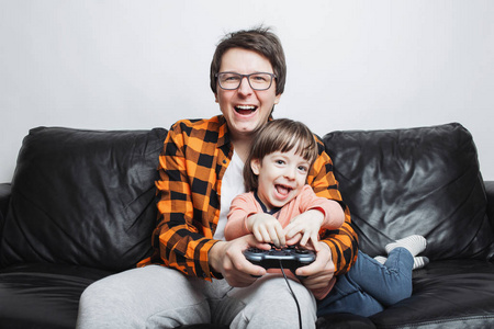 一个小帅哥和他爸爸坐在家里的沙发上，用操纵杆玩电子游戏。 爸爸和儿子在白色背景上玩得很开心。 他们快乐而微笑。