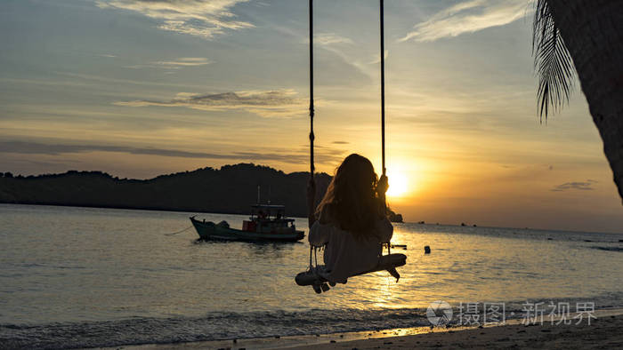 一个女孩的剪影,她坐在美丽的黄昏海滩上的普吉岛上的秋千上,寻找日落