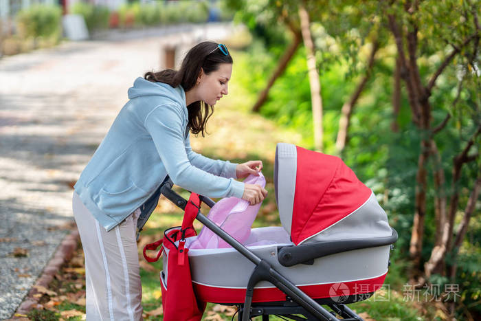 年轻的母亲看着的婴儿车在公园里散步