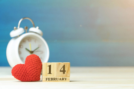 情人节的概念。 手工制作纱线红心旁边的木块日历设定在情人节日期2月14日在白色闹钟前的木制桌子和蓝色背景