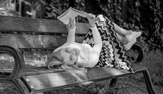 有趣的书。聪明又漂亮聪明的女士放松。女孩卧椅休闲与书, 绿色自然背景。妇女花闲暇与书。在板凳上放松的女孩在户外看书
