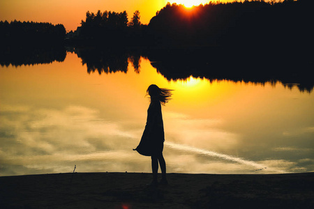 一个年轻美丽的女孩在夕阳的背景下在池塘的倒影中的剪影。