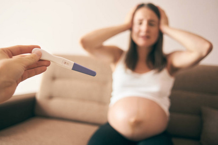 男性手中妊娠试验的选择性焦点和失望的年轻女性坐在沙发上，双手保持腹部