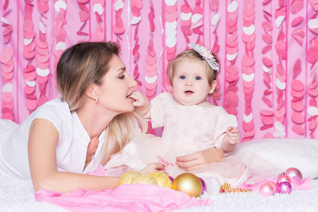 幸福的年轻母亲和她的女儿在柔软的毯子上的肖像，圣诞球和珠子在喜庆的粉红色内部。 妈妈和她的宝贝女儿一起玩得很开心。