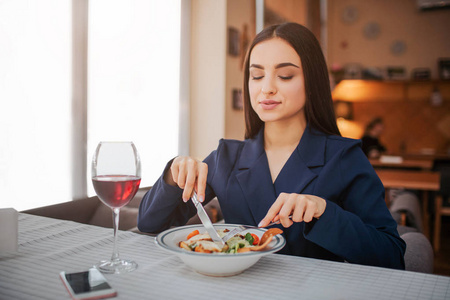骄傲善良的年轻女人坐在桌子旁，用叉子和刀吃沙拉。 她往下看。 还有一杯红酒摊。 躺在桌子上的电话