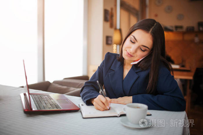 漂亮的年轻女子坐在桌旁通电话.她在笔记本上写字.女商人在餐馆工作.