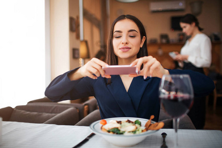 好年轻的女人在电话摄像机上拍沙拉碗的照片。 她看着它的屏幕。 除了一杯酒架