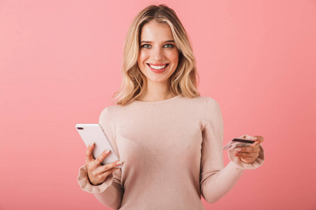 一位穿着毛衣的幸福年轻女子的肖像，她用手机在粉红色的背景下孤立地站着，显示着信用卡