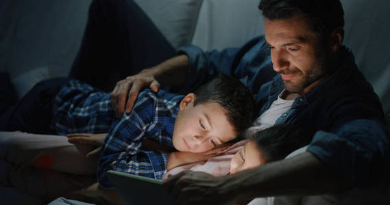 快乐父子的肖像，晚上在沙发上用平板电脑慢动作。 家庭娱乐教育技术的概念