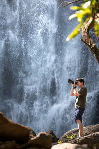 亚洲男子旅行者和摄影师使用数码相机在瀑布拍照。 旅行生活方式或自然摄影概念