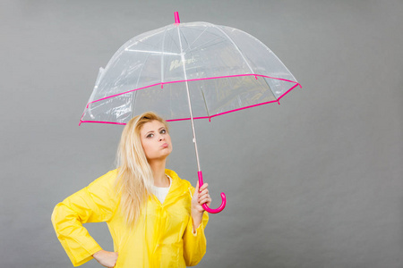悲伤无聊的金发女人穿着黄色雨衣，拿着透明的雨伞等着下雨。