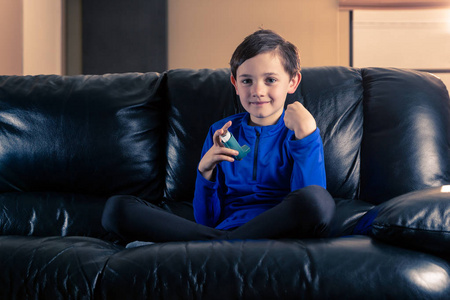 8岁男孩穿着带有哮喘吸入器的运动服，紧握拳头坐在沙发上。 健康疾病兴奋剂