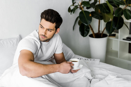 体贴的年轻人穿着睡衣，在床上拿着一杯咖啡
