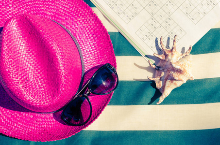 五颜六色的假日背景与粉红色稻草海滩帽子与太阳镜和数独在条纹日光浴床。 暑假海滩休闲活动在阳光下的概念。 平躺着。 上面的风景。 