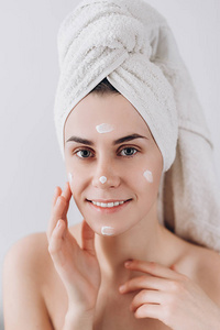 年轻快乐的女人用浴巾裹着，把奶油涂在脸上，用于护肤。 护肤概念。 令人惊叹的高加索女人，完美的健康，干净的皮肤。