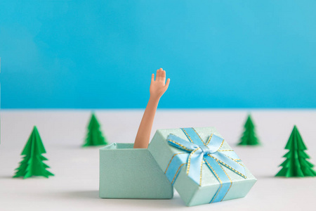 娃娃手臂从礼品盒和圣诞树制成的纸在蓝色背景下，最小的创意假日概念。