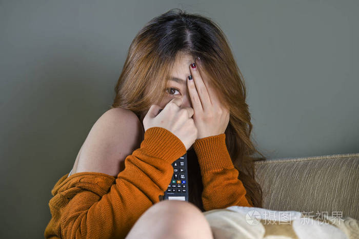 年轻,迷人,可爱,害怕的亚洲女人独自在家客厅沙发上看恐怖电影或电视