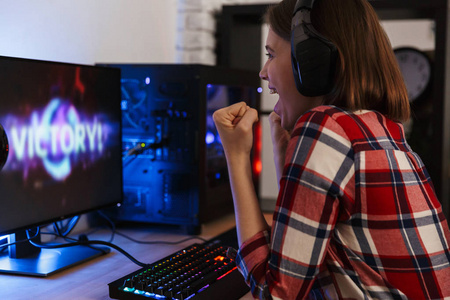 兴奋的女孩玩家坐在桌子旁玩电脑上的网络游戏，在室内庆祝胜利