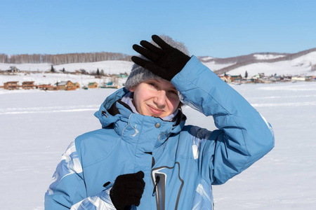 一个年轻的女孩滑雪者被一只来自太阳的手挡住了，站在村庄背景下的一个雪白的冰冻湖上。