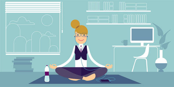 戴着耳机的戴着眼镜的瑜伽女孩坐在办公室里，坐在普拉提垫子上，摆出一个简单的苏哈萨纳姿势。 在她旁边，桌子上有一瓶水和一台电脑。