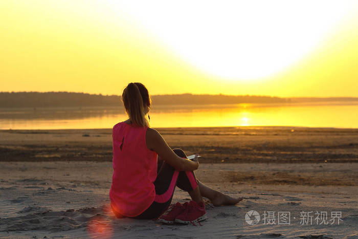 年轻漂亮的女孩穿着运动服坐在沙滩上,在海边享受橙