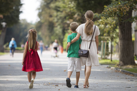 和儿子沿着阳光明媚的公园小巷,在模糊的人剪影和绿色的树木背景散步