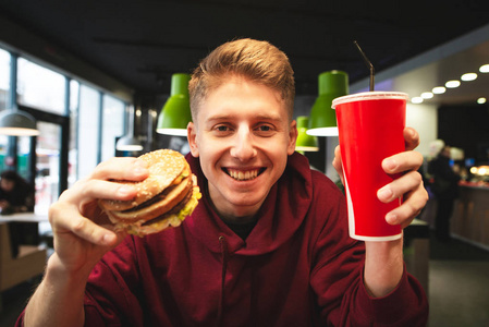 学生拿着一个汉堡和一杯可乐在一家快餐店的背景下。靠近一个微笑的年轻人的肖像，手里拿着一个汉堡和一个红玻璃，手里拿着一杯饮料，看着