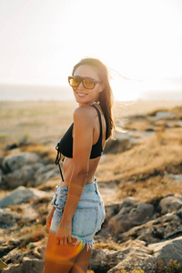 日落时海滩上戴太阳镜和牛仔裤的漂亮年轻女人的时尚肖像，复古风格。